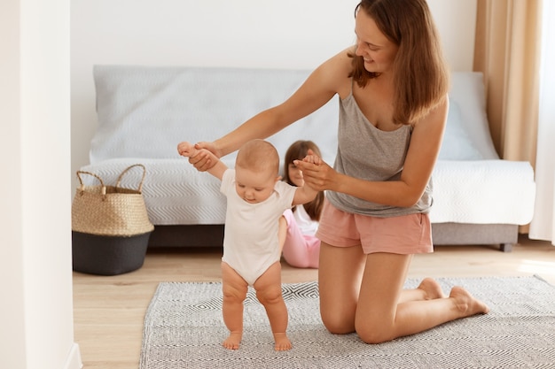 Ritratto di madre felice in piedi in ginocchio sul pavimento in soggiorno e insegnando al suo bambino ad andare, bambina che impara andare, infanzia felice.