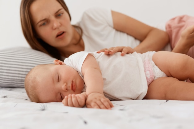 Ritratto di madre preoccupata spaventata guardando la sua piccola figlia addormentata, toccando il bambino, femmina con i capelli scuri che indossa una maglietta bianca in stile casual, maternità.