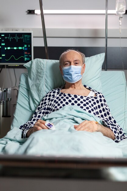 Ritratto di malato uomo anziano paziente con maschera chirurgica in appoggio nel letto di ospedale, con flebo iv attaccato a portata di mano con ossimetro attaccato al dito