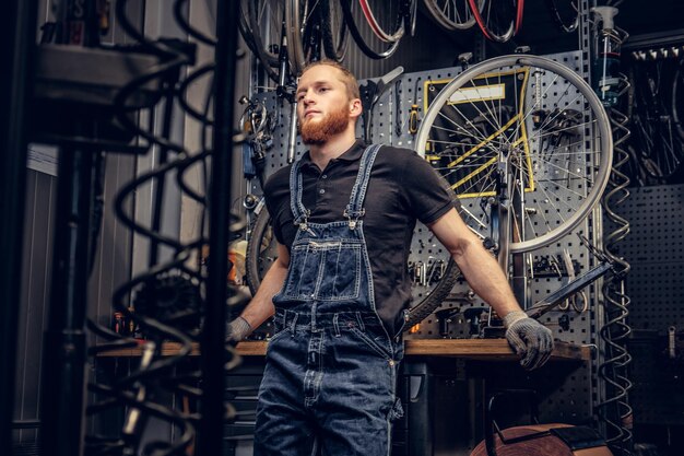 Ritratto di meccanico di biciclette barbuto testa rossa in un'officina con parti di biciclette e ruota su uno sfondo.