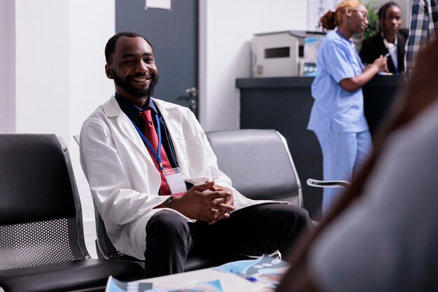 Ritratto di medico afroamericano nella hall, seduto nei sedili della sala d'attesa prima di avere un appuntamento per la consultazione medica con i pazienti. Medico generico che lavora sull'assistenza sanitaria nel centro.