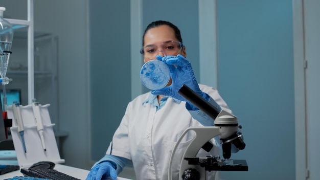 Ritratto di medico di chimica che fa indagini scientifiche in laboratorio con apparecchiature chimiche. Donna che utilizza la capsula di Petri per la progettazione del DNA su computer, con microscopio e vacutainer