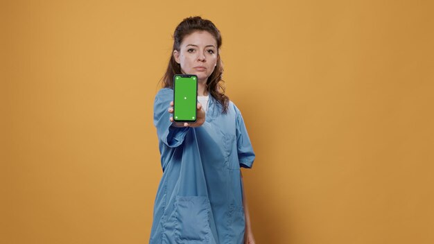 Ritratto di medico professionista che tiene smartphone e manda messaggi di testo e mostra il design del mockup dello schermo verde alla fotocamera in studio. Medico in uniforme ospedaliera utilizzando un dispositivo touchscreen che presenta lo spazio della copia.