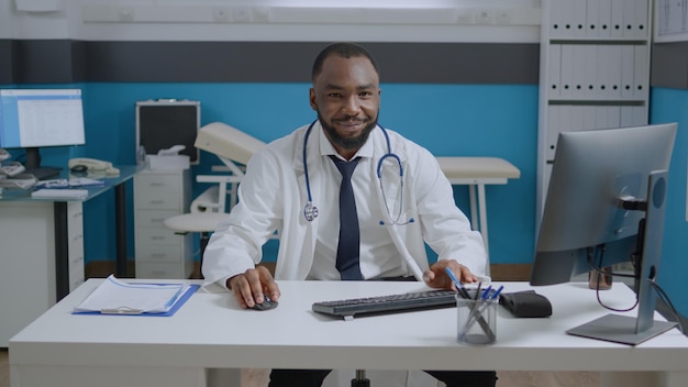 Ritratto di medico specialista afroamericano seduto alla scrivania digitando trattamento farmacologico analizzando il rapporto diagnostico di malattia. Uomo del medico che lavora con esperienza medica nell'ufficio dell'ospedale