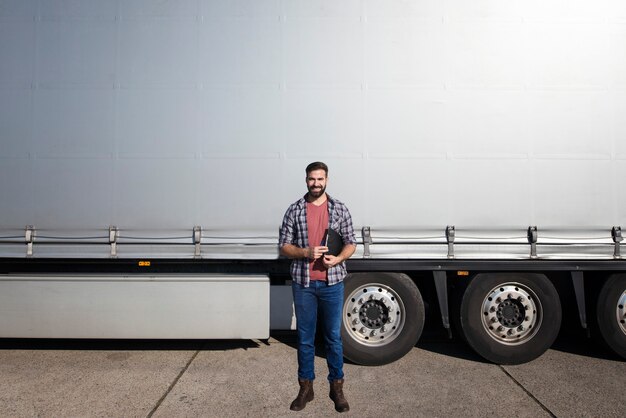 Ritratto di mezza età barbuto camionista in piedi davanti al rimorchio del camion contro il telone lucido grigio