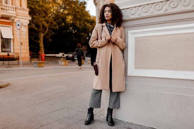 Ritratto di moda all'aperto di glamour sensuale giovane elegante signora nera che indossa un vestito alla moda autunno, maglione di velluto grigio e cappotto di lana beige.