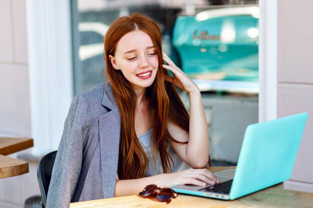 Ritratto di moda città all'aperto di giovane donna di affari che lavora al caffè sulla terrazza al giorno pieno di sole, vestito elegante casual, dettagli di menta, utilizzando il suo computer portatile, pausa caffè, concetto di affari.