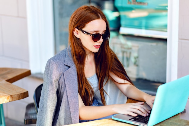 Ritratto di moda città all'aperto di giovane donna di affari che lavora al caffè sulla terrazza al giorno pieno di sole, vestito elegante casual, dettagli di menta, utilizzando il suo computer portatile, pausa caffè, concetto di affari.