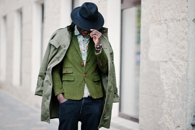 Ritratto di moda di uomo afroamericano nero su giacca di velluto verde cappello nero e mantello sulle spalle camminando per le strade della casa di sfondo della città con molte finestre