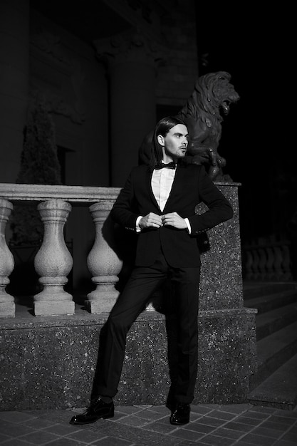 Ritratto di moda elegante giovane dai capelli lunghi. Modello maschio attraente e bello in abito nero con i baffi in strada di notte