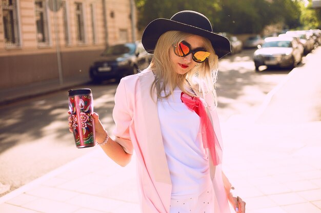 Ritratto di moda stile di vita soleggiato estate di giovane donna alla moda hipster che cammina per strada, indossando abiti alla moda carino, bevendo latte caldo