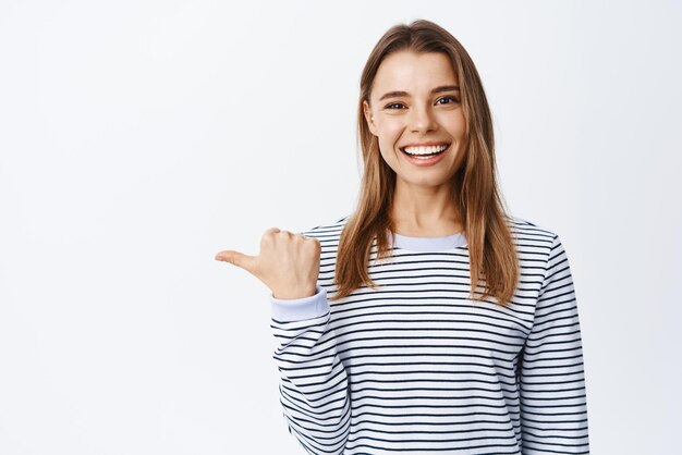 Ritratto di modello femminile allegro con denti bianchi che puntano a sinistra invitando a controllare o guardare il logo che mostra lo sfondo bianco della pubblicità