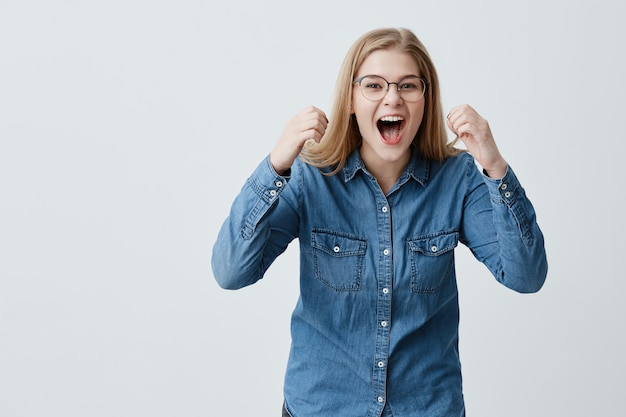 Ritratto di modello femminile eccitato e felicissimo con capelli biondi, occhiali, in camicia di jeans, stringe i pugni con piacere, grida di felicità, celebra la vittoria, ha un grande trionfo. Concetto di successo