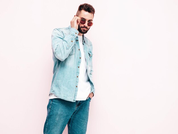 Ritratto di modello lambersexual bello e sicuro di sé hipster uomo vestito con giacca e jeans Moda maschile in posa vicino al muro rosa in studio in occhiali da sole