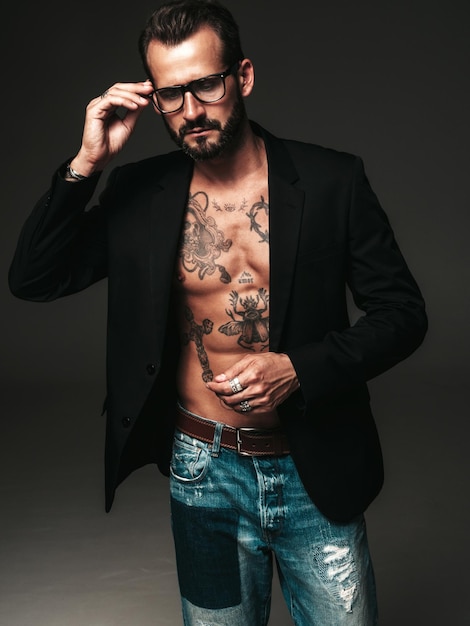 Ritratto di modello lambersexual bello fiducioso elegante hipster Uomo moderno sexy vestito con giacca nera Torso nudo con tatuaggiUomo di moda in posa in studio su sfondo scuro In occhiali