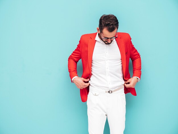 Ritratto di modello lambersexual bello fiducioso elegante hipster Uomo moderno sexy vestito con un elegante abito rosso Moda maschile in posa in studio vicino al muro blu in occhiali da sole