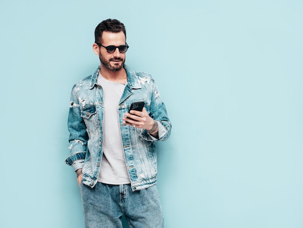 Ritratto di modello lambersexual bello fiducioso elegante hipster Uomo sexy vestito con giacca di jeans Moda maschile isolato sulla parete blu in studio In occhiali da sole