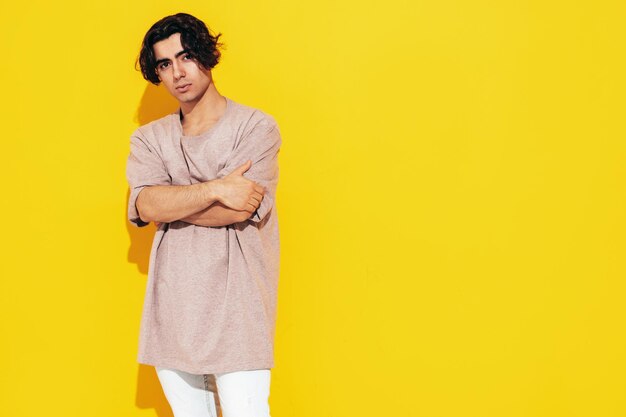 Ritratto di modello lambersexual bello fiducioso elegante hipster Uomo sexy vestito con maglietta e jeans di grandi dimensioni Moda maschio isolato in studio in posa vicino al muro giallo