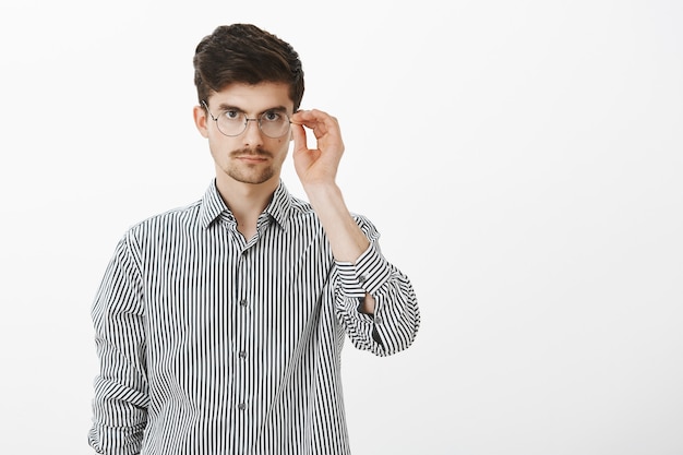 Ritratto di modello maschio nerd dall'aspetto serio con barba e baffi, che tiene il bordo degli occhiali, guardando concentrato, ascoltando attentamente il capo durante la riunione, pronto per iniziare a lavorare sul progetto