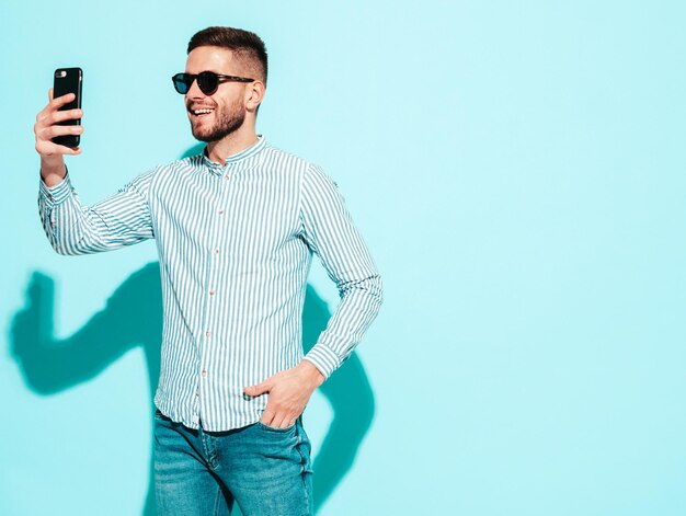 Ritratto di modello sorridente bello Uomo alla moda sexy vestito in camicia e jeans Moda uomo hipster in posa vicino al muro blu in studio Allegro e felice Scattare foto selfie