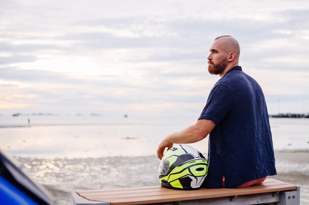 Ritratto di motociclista maschio con casco giallo seduto su una panchina sul lungomare in Thailandia al tramonto