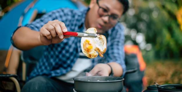 Ritratto di occhiali da uomo viaggiatore asiatico che friggono un gustoso uovo fritto in una padella calda al campeggio Cucina all'aperto concetto di stile di vita in campeggio in viaggio