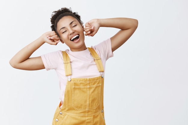 Ritratto di pigra affascinante studentessa afroamericana in elegante tuta gialla, che si estende con le mani alzate, sbadigliando e sorridendo ampiamente, sensazione di sonnolenza dopo il pisolino