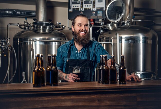 Ritratto di produttore maschio hipster tatuato e barbuto che presenta un microbirrificio di birra.