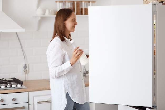 Ritratto di profilo di attraente donna dai capelli scuri che indossa una camicia bianca, guardando sorridente all'interno del frigorifero con emozioni positive, tenendo il piatto in mano, posando con set da cucina sullo sfondo.