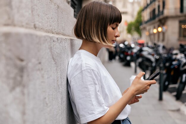 Ritratto di profilo di elegante ragazza dai capelli corti in maglietta bianca smartphone a scorrimento in città Ragazza caucasica alla moda con smartphone in posa all'aperto con un sorriso