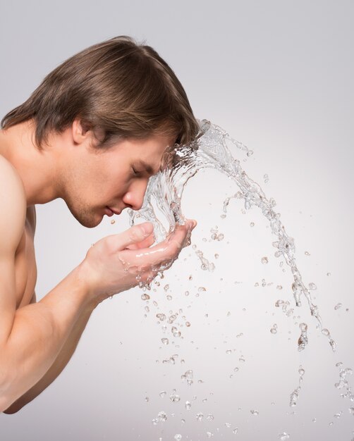 Ritratto di profilo di un uomo bello che si lava il viso con acqua sul muro grigio.