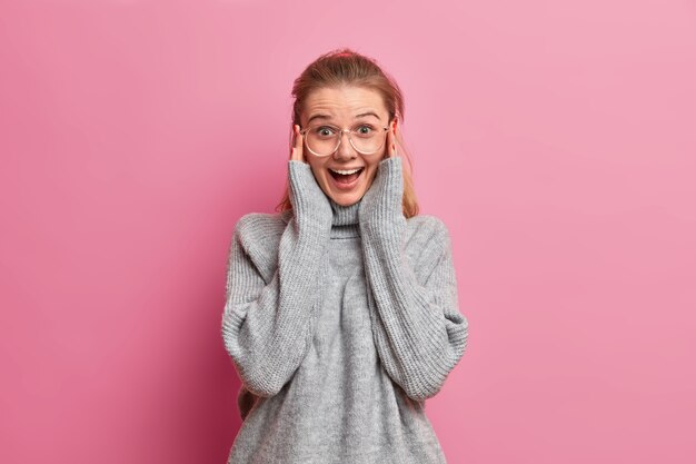 Ritratto di ragazza abbastanza allegra indossa grandi occhiali da vista e maglione oversize, ridacchia positivamente, guarda uno spettacolo divertente