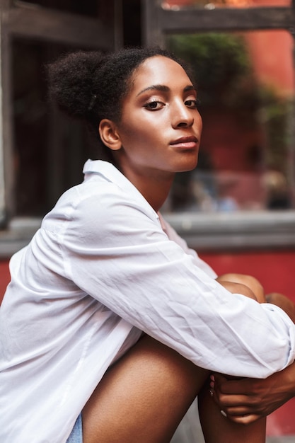 Ritratto di ragazza africana premurosa con capelli ricci scuri in camicia bianca che guarda sognante nella fotocamera isolata