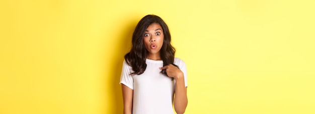 Ritratto di ragazza afroamericana sorpresa che punta il dito contro se stessa in piedi su sfondo giallo