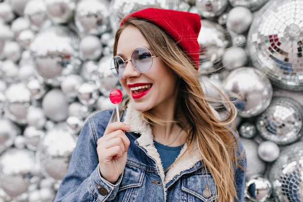 Ritratto di ragazza alla moda in giacca di jeans, mangiare caramelle e distogliere lo sguardo. Meravigliosa signora europea con cappello rosso e occhiali rotondi in posa con lecca-lecca sul muro lucido.