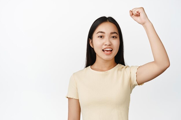 Ritratto di ragazza asiatica allegra che alza la mano per protestare e cantare guardando incoraggiata in piedi su sfondo bianco