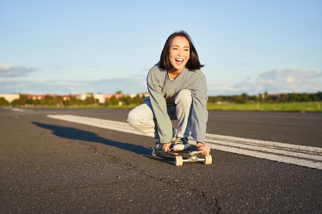 Ritratto di ragazza asiatica felice spensierata che pattina in sella a skateboard e ride godendosi la giornata di sole