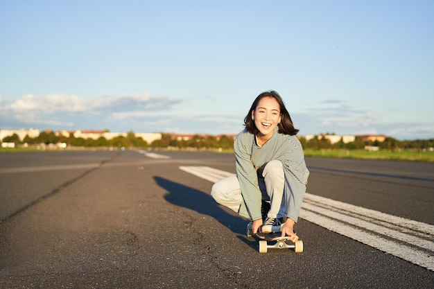 Ritratto di ragazza asiatica felice spensierata che pattina in sella a skateboard e ride godendosi la giornata di sole