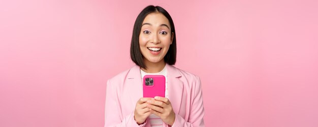 Ritratto di ragazza asiatica in tuta con smartphone sorridente e guardando felice in piedi su sfondo rosa studio