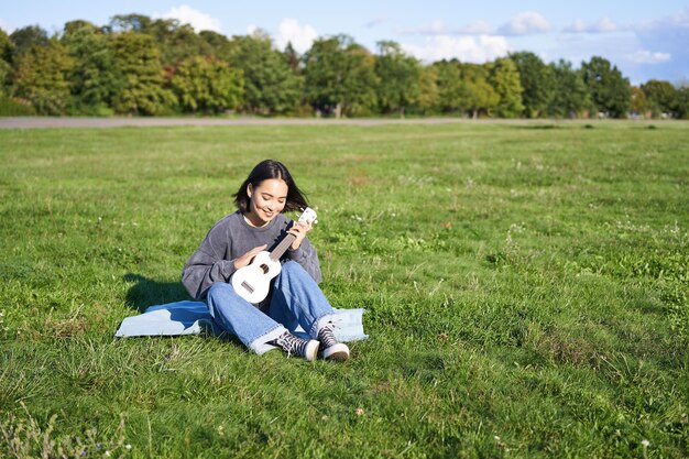 Ritratto di ragazza asiatica seduta da sola nel parco che suona ukulele e canta godendosi il tempo libero