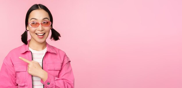 Ritratto di ragazza asiatica sorridente in occhiali da sole vestito elegante che punta il dito a sinistra mostrando banner pubblicitario in piedi su sfondo rosa Spazio di copia