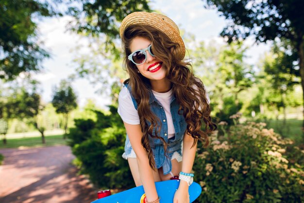 Ritratto di ragazza attraente con capelli ricci lunghi in cappello in posa con lo skateboard nel parco estivo. Indossa farsetto di jeans, occhiali da sole. Lei sta sorridendo.