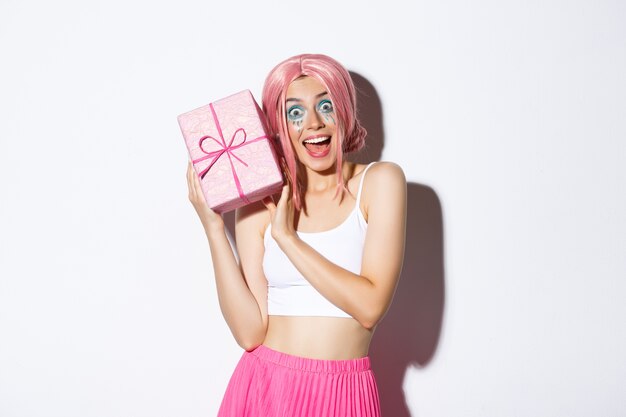 Ritratto di ragazza attraente sorpresa che sembra eccitata, riceve un regalo per il compleanno, indossa una parrucca rosa, in piedi.