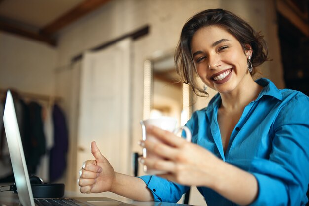 Ritratto di ragazza bella studentessa allegra in vestito blu che sorride ampiamente, divertendosi mentre naviga in Internet sul computer portatile