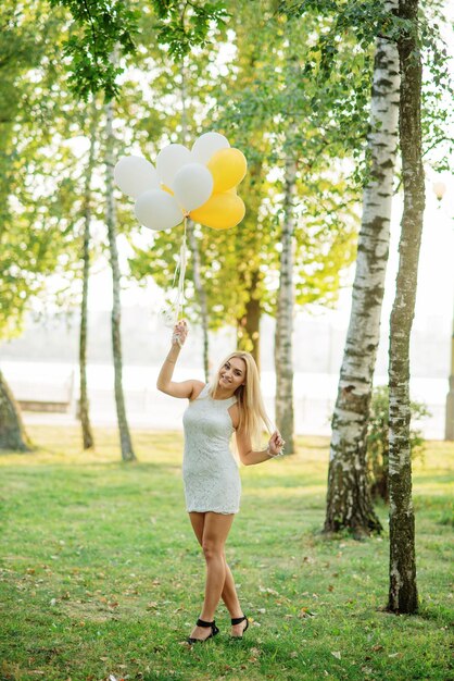 Ritratto di ragazza bionda che indossa un abito bianco con palloncini a portata di mano contro il parco alla festa di addio al nubilato
