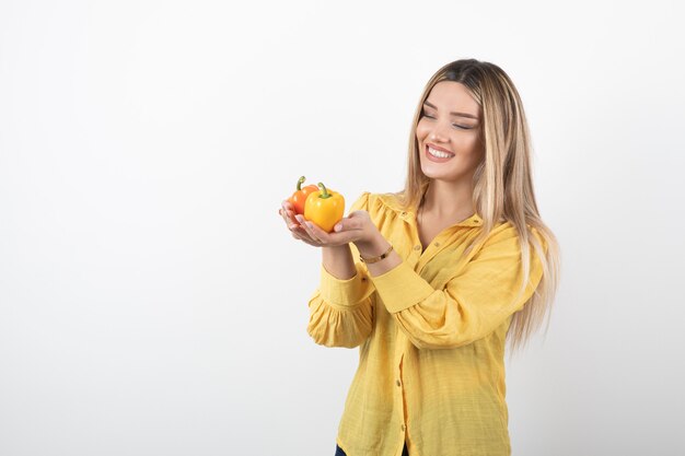 Ritratto di ragazza bionda che regala peperoni colorati sul muro bianco.