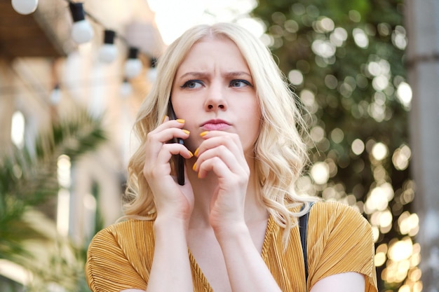 Ritratto di ragazza bionda seria sconvolta che distoglie premurosamente lo sguardo mentre parla al cellulare all'aperto