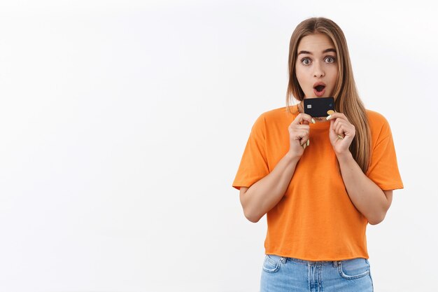 Ritratto di ragazza bionda sorpresa ed eccitata in maglietta arancione non vedo l'ora di sprecare tutti i suoi soldi in un nuovo vestito per le vacanze estive, in possesso di carta di credito, fissando la telecamera impressionata, facendo shopping online