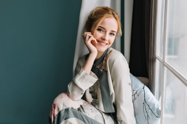 Ritratto di ragazza bionda sorridente e felice accanto alla finestra, rilassante al mattino, divertendosi a casa. È vestita con un bel pigiama di seta.