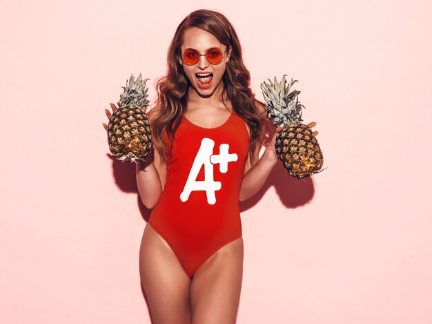 Ritratto di ragazza bruna sorridente in abiti da bagno rosso estate e occhiali da sole rotondi. Donna sexy con ananas fresco. Posa positiva del modello